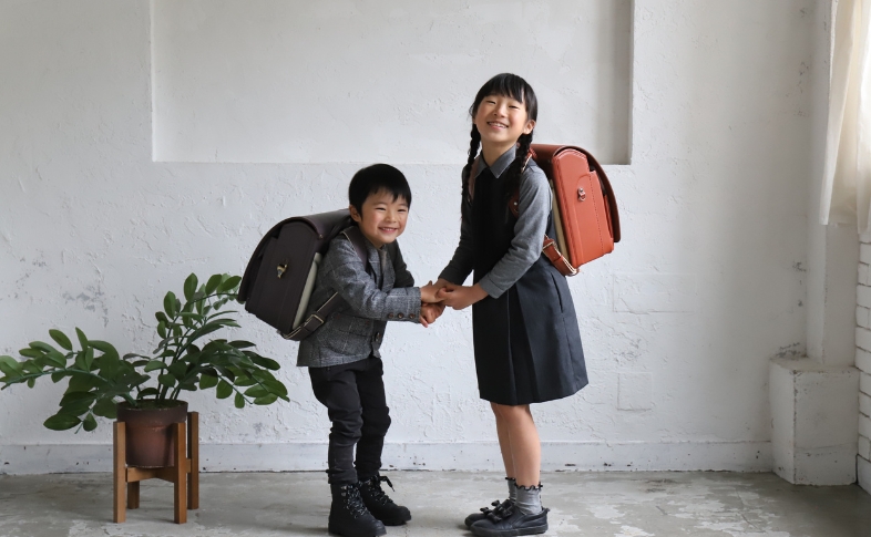 手を取り合った小学生の男の子と女の子が、楽しそうな様子でこちらに笑顔を見せている。男の子はラベンダーのランドセル、女の子はオレンジのランドセルを背負っている。