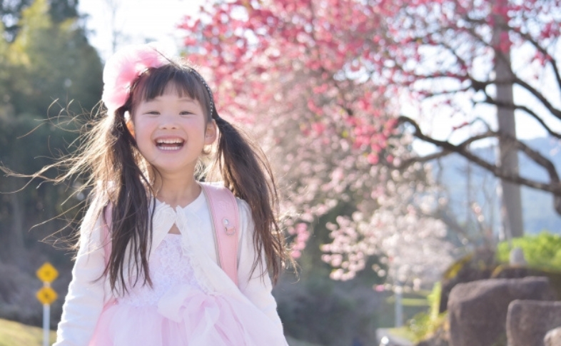 桜を背景に、ピンクのワンピースと白のボレロを着用している笑顔の女の子